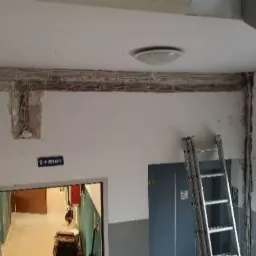 Instalacja elektryczna w korytarzu, widoczne kable ułożone w bruzdach pod sufitem, drabina oparta o ścianę, surowy stan wykończenia.