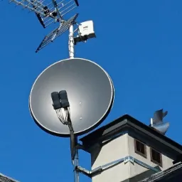 Antena telewizyjna Yagi-Uda i antena satelitarna zamontowane na kominie budynku z szarą dachówką na tle błękitnego nieba.