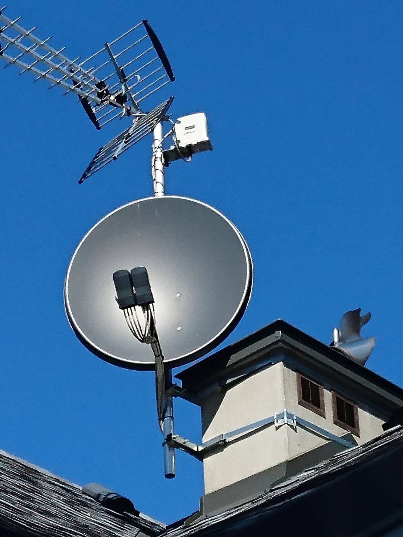 Antena telewizyjna Yagi-Uda i antena satelitarna zamontowane na kominie budynku z szarą dachówką na tle błękitnego nieba.