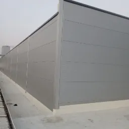 Szara ściana hali przemysłowej wykonana z poziomych płyt warstwowych, widok z zewnątrz, z widocznym fragmentem dachu i rynny.