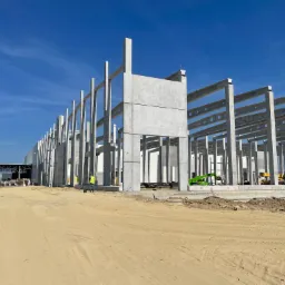 Konstrukcja hali w trakcie budowy: szkielet z prefabrykatów betonowych, widoczny pracownik w żółtej kamizelce, żurawie i maszyny budowlane na piaszczystym terenie.