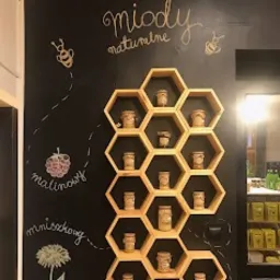 Ściana z czarną tablicą, na której kredą narysowane są pszczoły, malina i mniszek lekarski oraz napis 'miody naturalne'. Na tablicy umieszczony drewniany regał w kształcie plastra miodu, wypełniony...