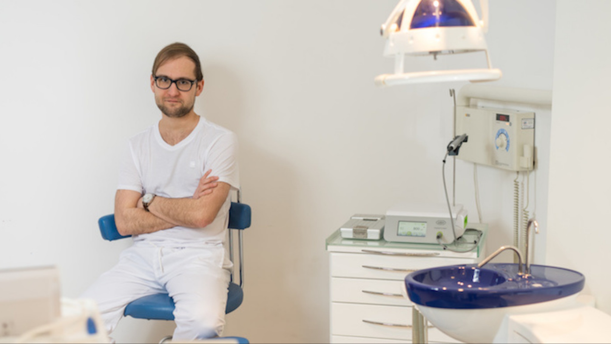 Lekarz w białym stroju siedzi na niebieskim krześle w gabinecie, w tle widoczny sprzęt dentystyczny i lampa zabiegowa.