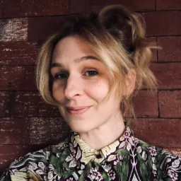 Portret uśmiechniętej kobiety z blond włosami upiętymi w kok, ubrana w koszulę w kwiaty na tle ceglanej ściany.