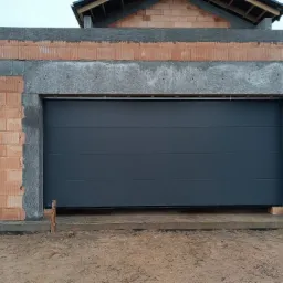 Szara segmentowa brama garażowa w surowym stanie, widoczna w trakcie montażu w nowo budowanym garażu z cegły, z widocznym betonowym nadprożem i częściowo otynkowanymi ścianami.