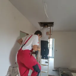 Dwóch monterów na drabinach, jeden wierci w suficie podwieszanym, drugi wchodzi do otworu rewizyjnego, widoczne narzędzia i elementy konstrukcji.