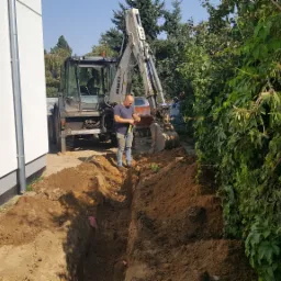 Operator koparki mierzy głębokość wykopu przy budynku jednorodzinnym, widoczne ślady pracy maszyny ziemnej.