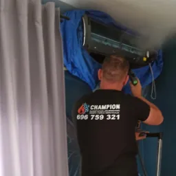 Mężczyzna w czarnej koszulce z logo firmy Champion, stojący na drabinie i używający narzędzia do czyszczenia klimatyzatora w pomieszczeniu z zasłonami i niebieską ścianą.
