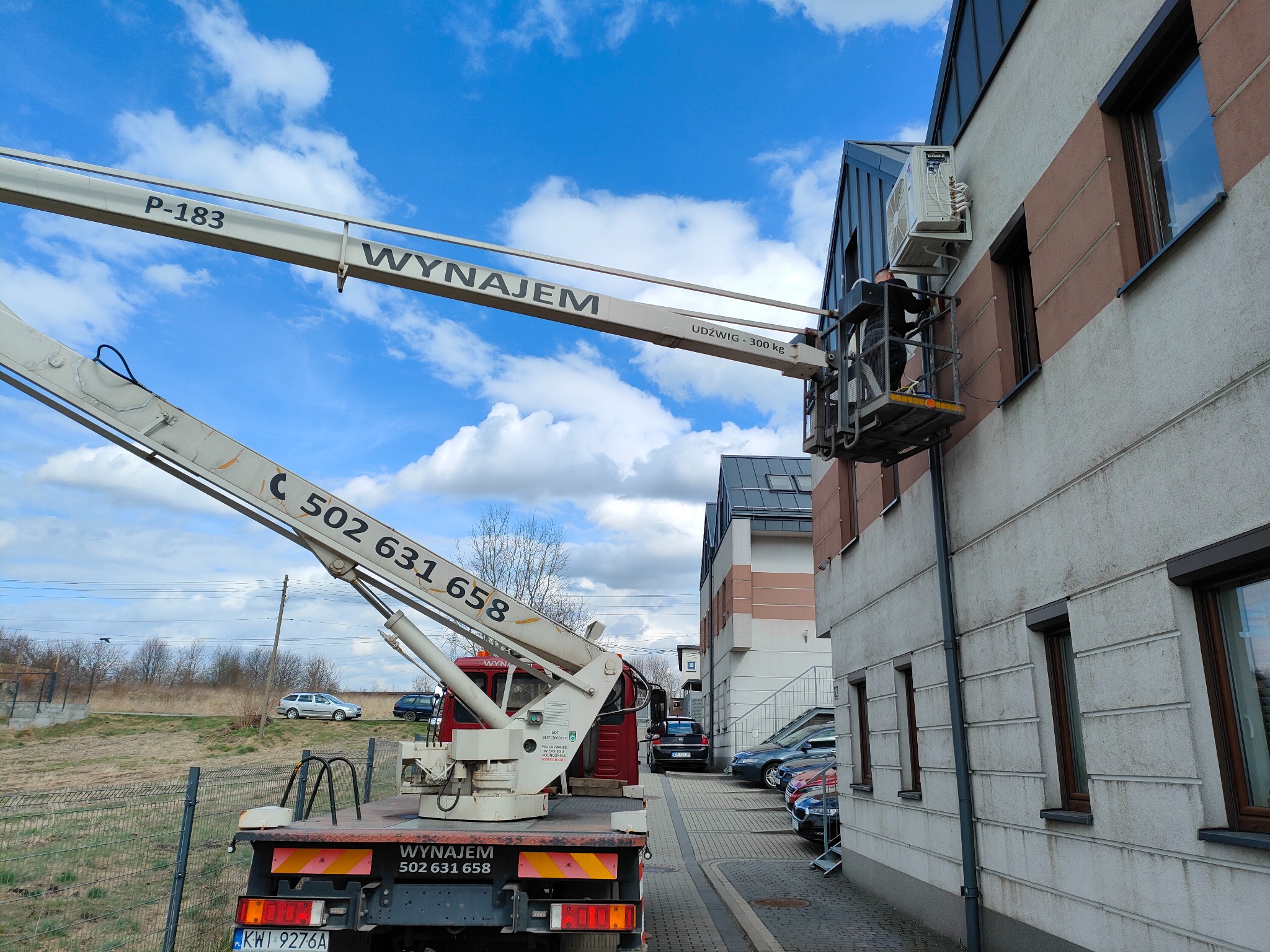 Montaż klimatyzacji na elewacji budynku z użyciem podnośnika koszowego. Widoczny pracownik w koszu, jednostka zewnętrzna klimatyzatora i fragment elewacji.