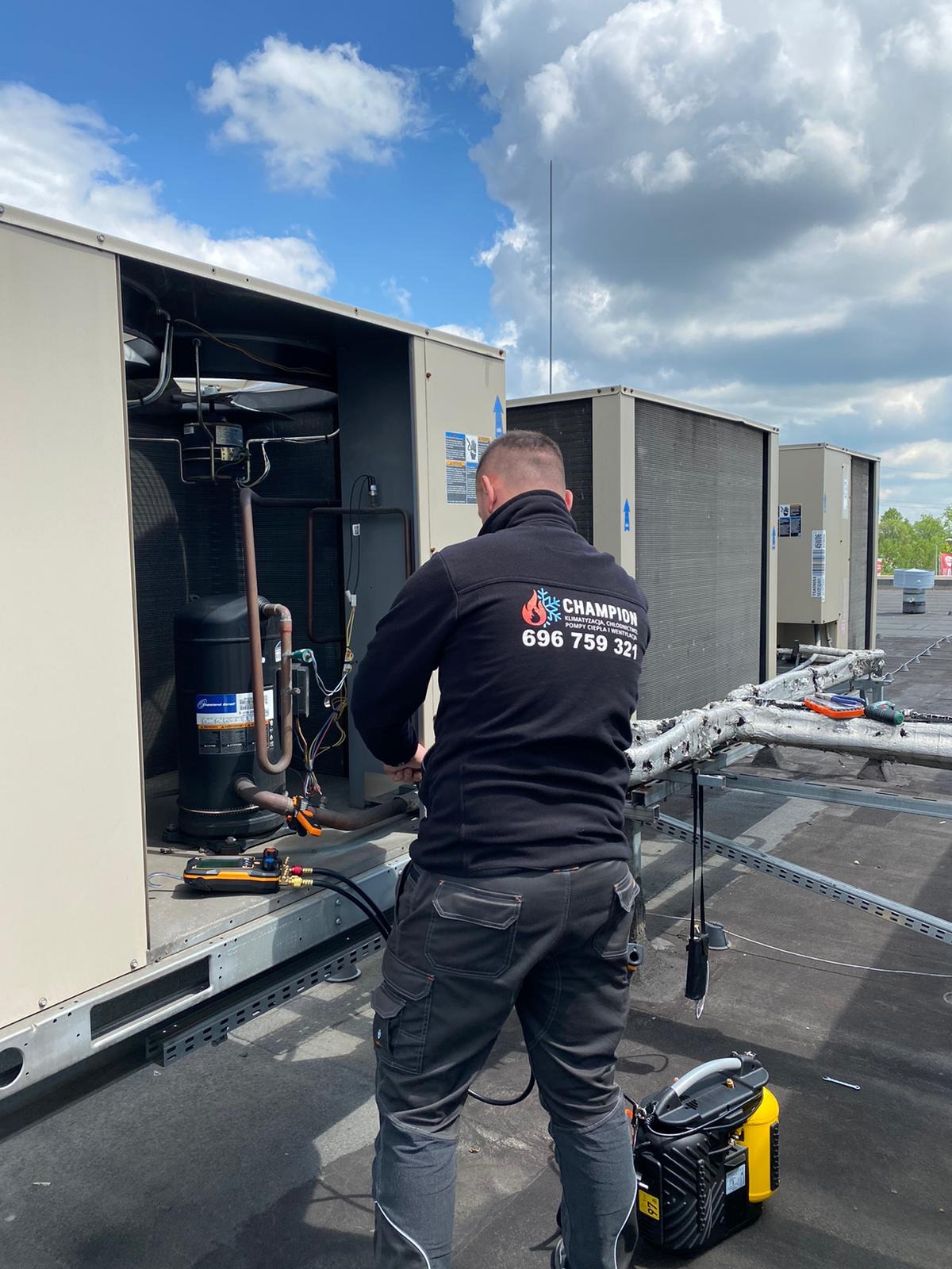 Serwisant w czarnej bluzie z logo firmy Champion naprawia klimatyzację na dachu budynku. Widoczne narzędzia i agregaty klimatyzacyjne. Błękitne niebo z chmurami w tle.