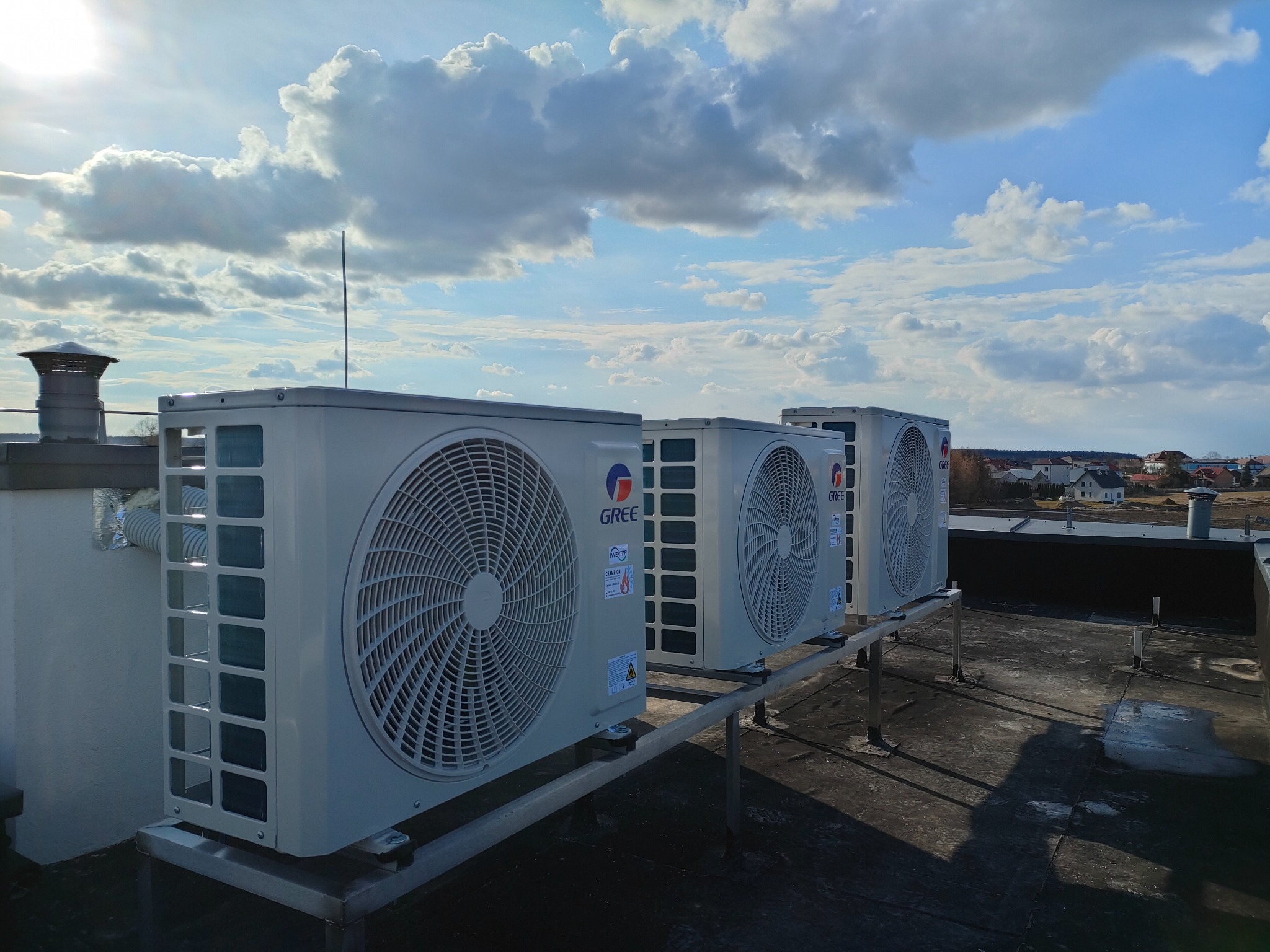 Trzy klimatyzatory Gree na dachu budynku w słoneczny dzień. Widoczne jednostki zewnętrzne, metalowa konstrukcja wsporcza i fragment panoramy miasta w tle.