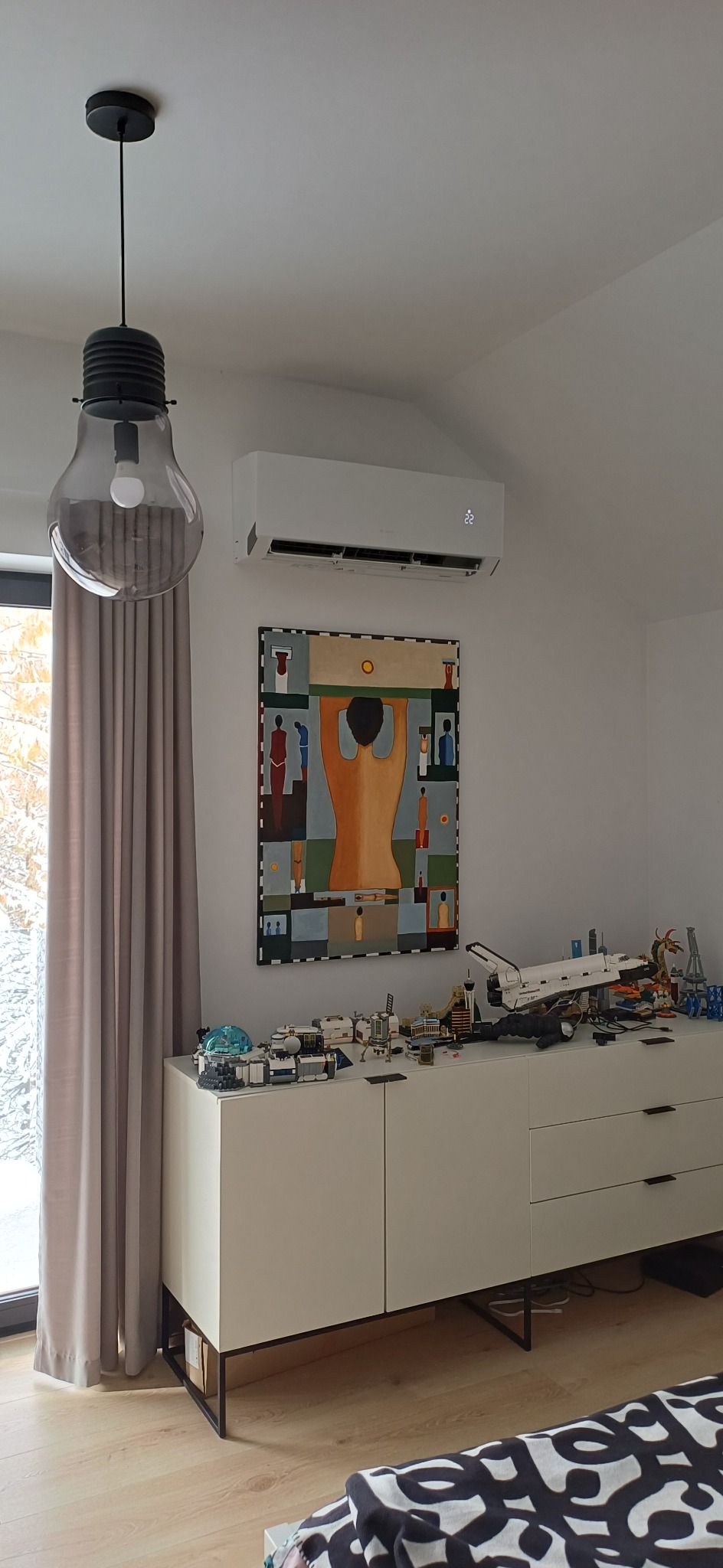 Biała klimatyzacja ścienna w nowoczesnym wnętrzu z obrazem i designerską lampą. Widoczny fragment okna i zasłony. Na komodzie zabawki.