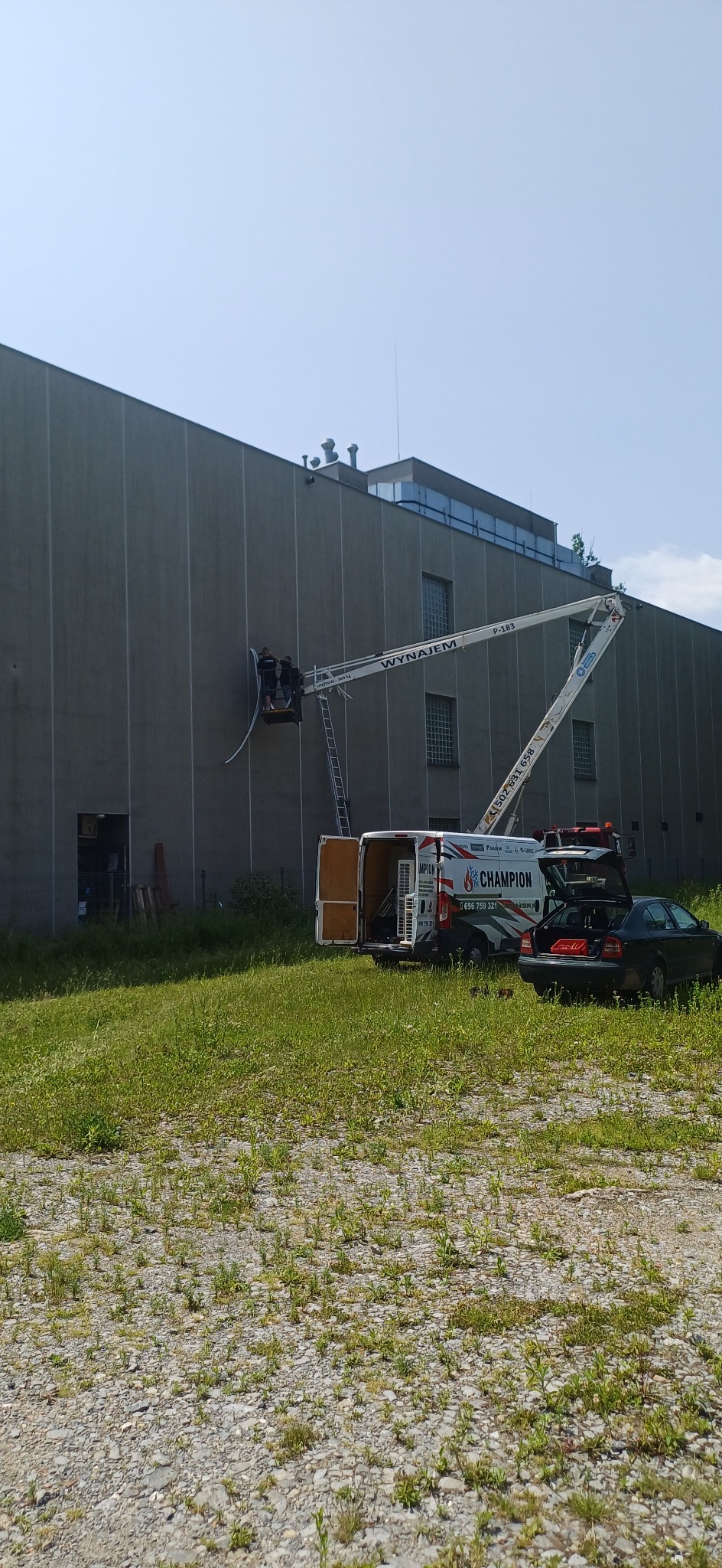 Montaż klimatyzacji na wysokości przy użyciu podnośnika koszowego. Widoczny budynek przemysłowy, trawa i samochód dostawczy z logo firmy Champion.