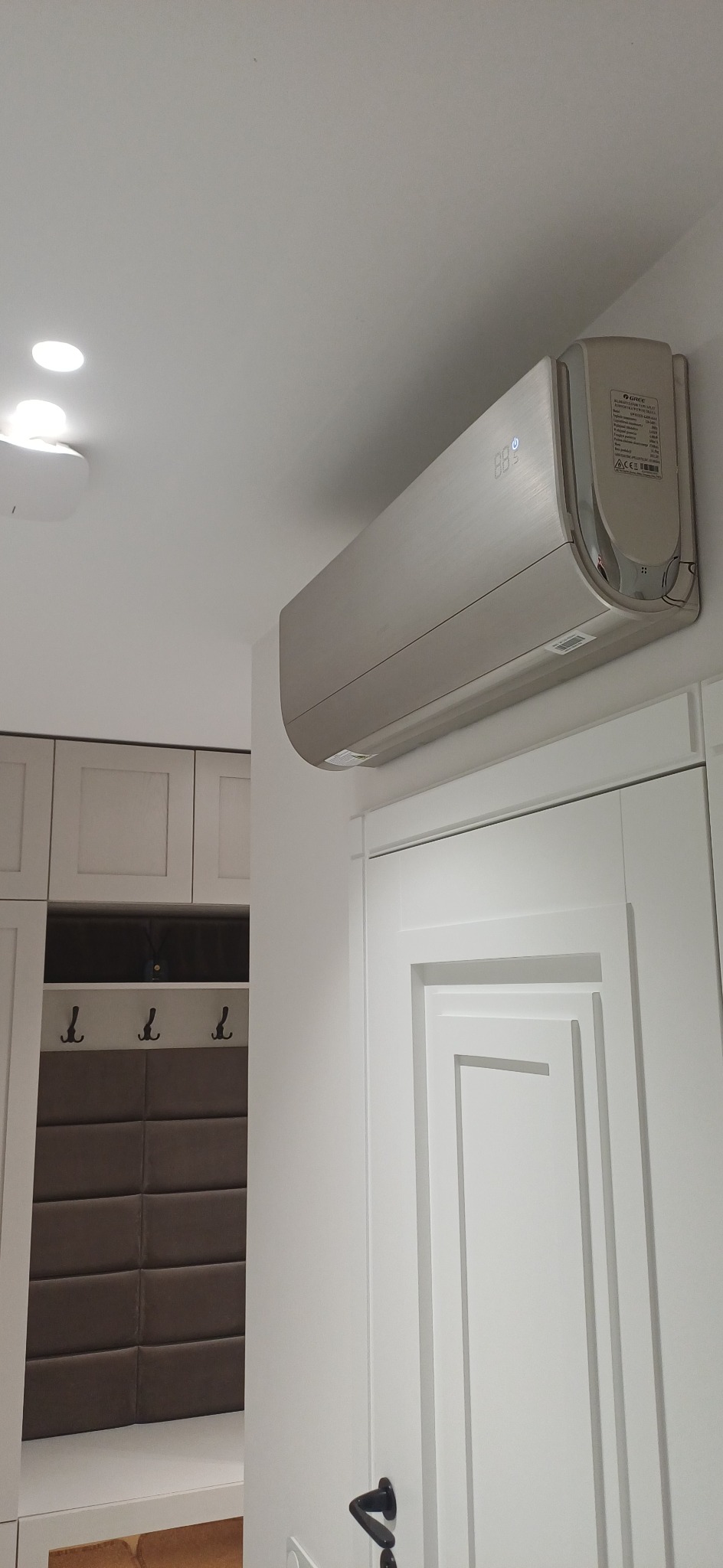 Nowoczesny, srebrny klimatyzator ścienny marki Gree zamontowany nad białą, frezowaną futryną drzwiową w jasnym wnętrzu. Widoczny fragment szafy z wieszakami.
