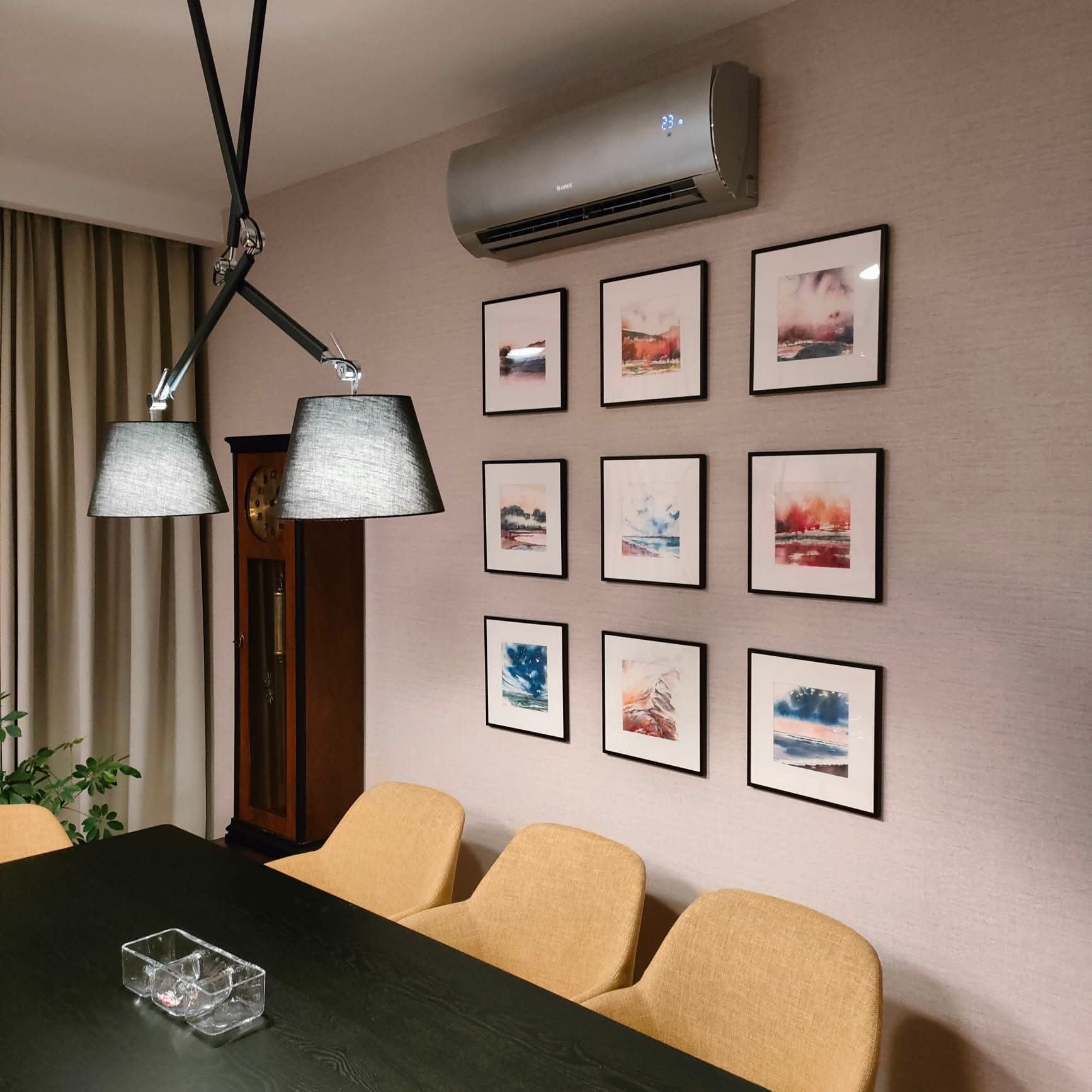 Szary klimatyzator ścienny Gree włączony, z wyświetlaczem temperatury 23°C, zawieszony nad galerią obrazów w czarnych ramkach w jadalni z zegarem i lampą.