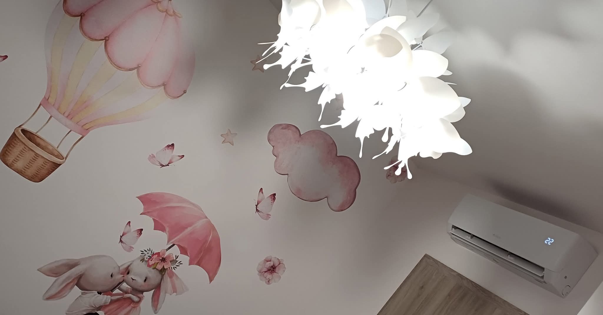 Biała klimatyzacja na suficie pokoju dziecięcego z dekoracyjnymi naklejkami: balon, chmurki, motyle i króliki. Nowoczesna lampa sufitowa.