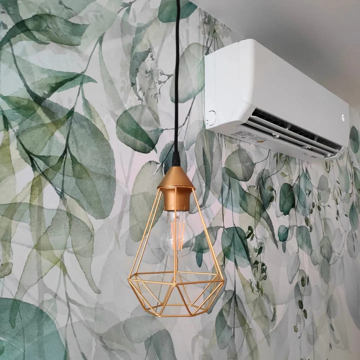 Biała klimatyzacja ścienna w pokoju z tapetą w liście, obok złota lampa wisząca. Nowoczesny design wnętrza, klimatyzator zamontowany na ścianie.