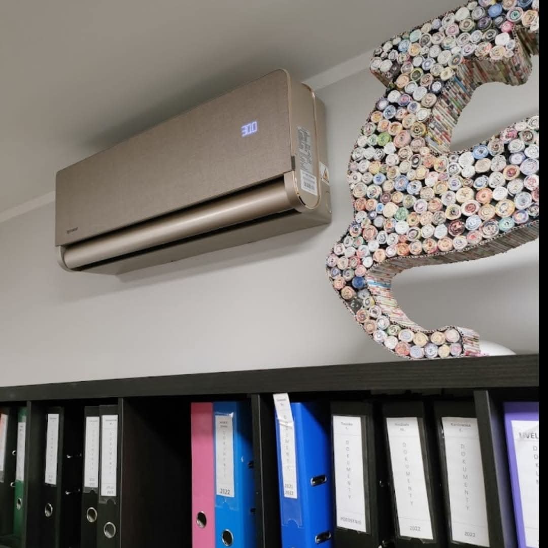 Beżowy klimatyzator ścienny z wyświetlaczem temperatury, zamontowany nad czarnym regałem z segregatorami. Obok dekoracja ścienna z rolek papieru.