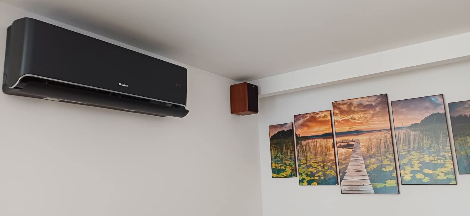 Czarny klimatyzator Gree na ścianie w pokoju z obrazami jeziora o zachodzie słońca. Widoczny głośnik na ścianie. Nowoczesny design i estetyczny montaż.