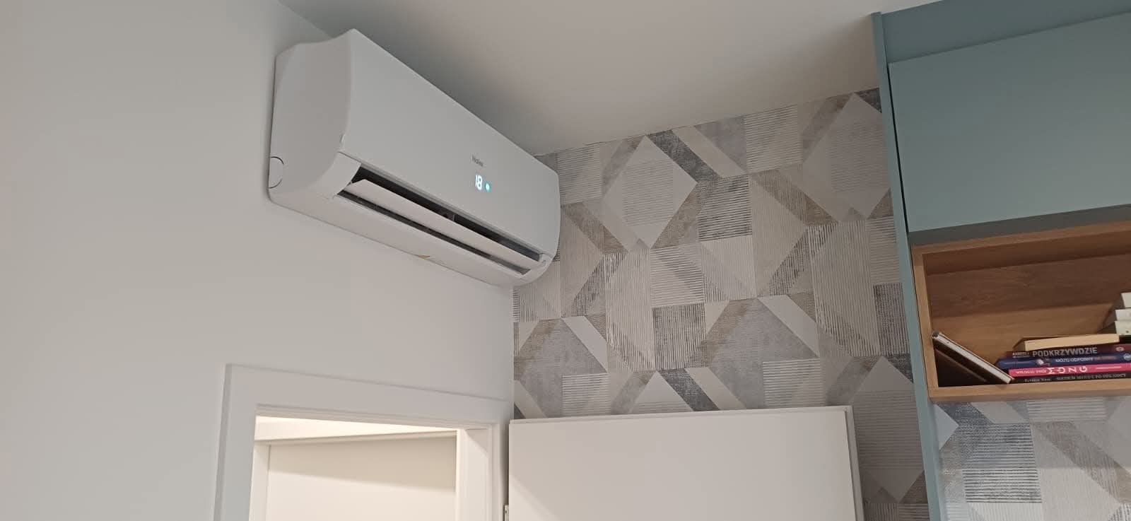 Biały klimatyzator Haier zamontowany na ścianie z geometryczną tapetą w pokoju. Widoczny fragment drzwi i regału z książkami. Nowoczesny design.