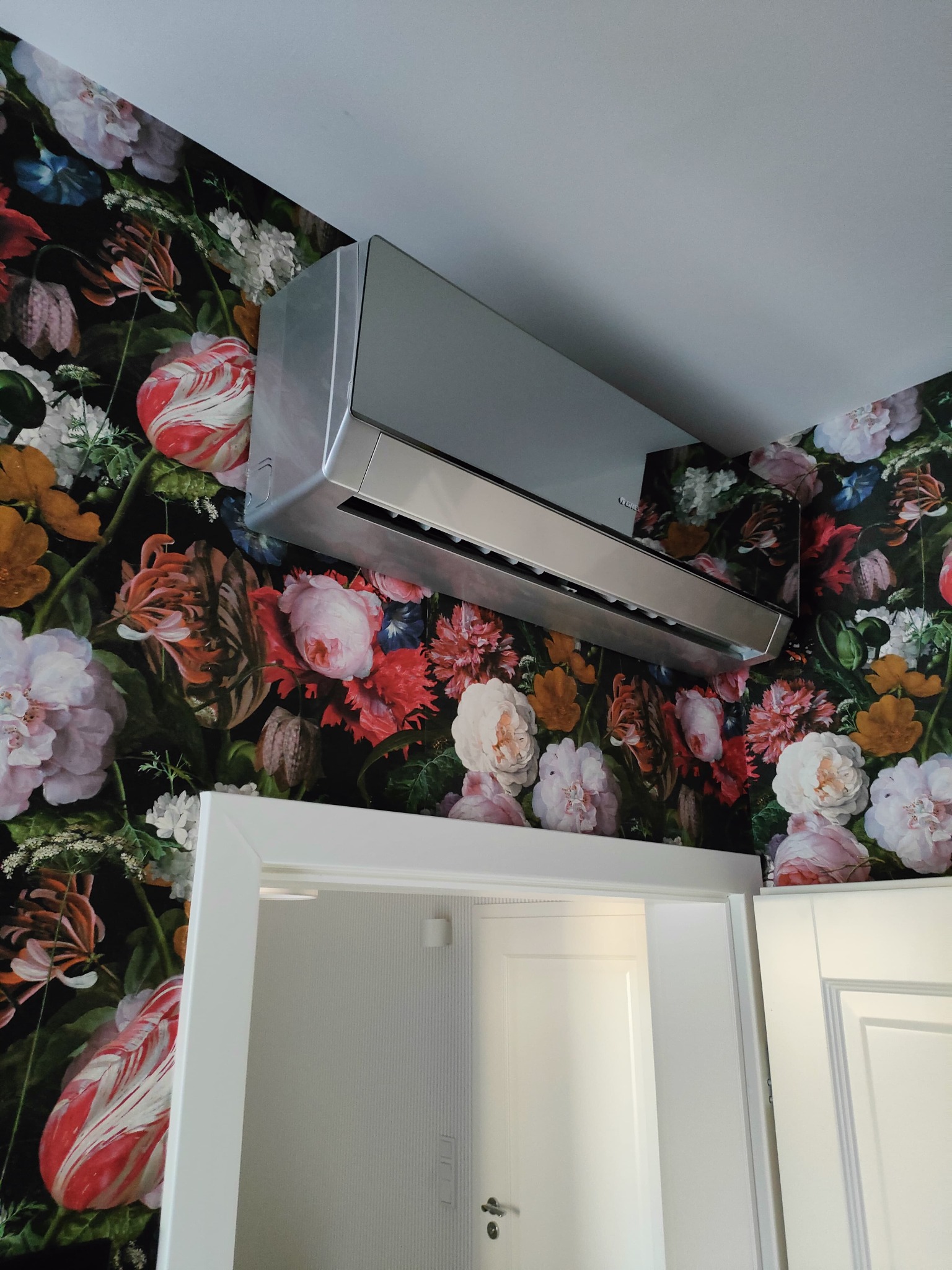 Srebrny klimatyzator na ścianie z motywem kwiatowym, widoczny nad otwartymi, białymi drzwiami. Nowoczesny design wkomponowany w dekoracyjne wnętrze.