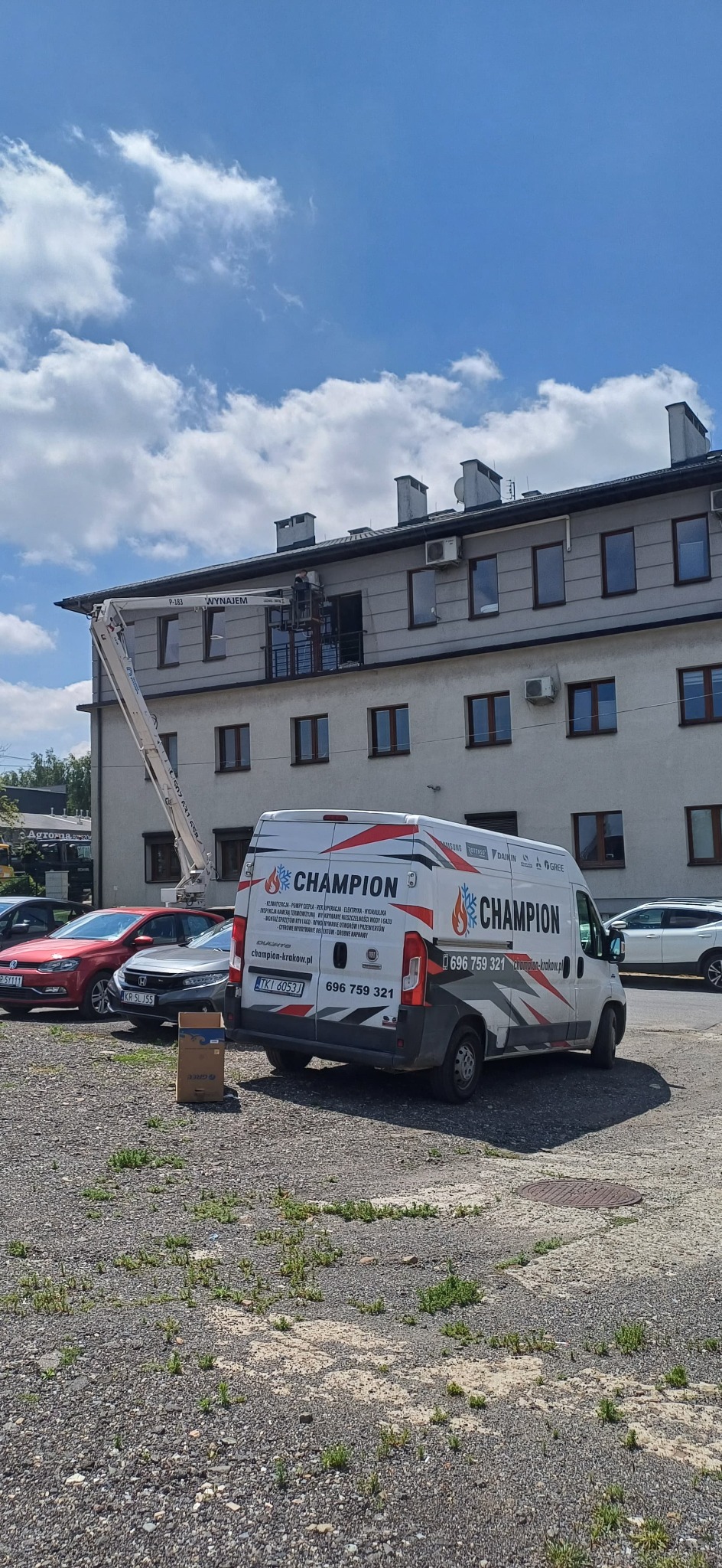 Montaż klimatyzacji w Krakowie z podnośnika. Na dachu budynku widoczny pracownik. Obok zaparkowany firmowy van z logo Champion i numerem telefonu.