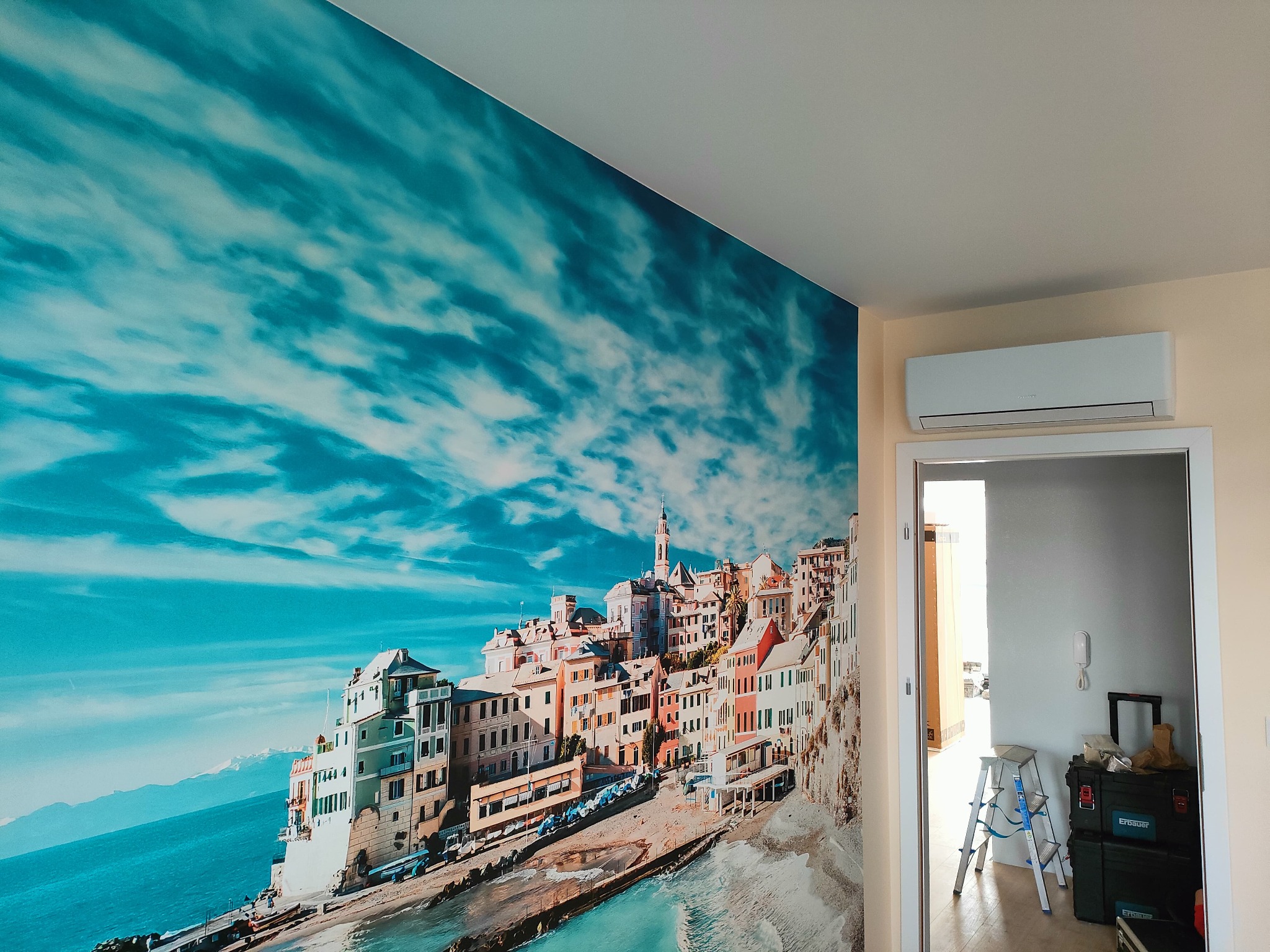 Biała klimatyzacja ścienna w pokoju z fototapetą przedstawiającą włoskie miasteczko nad morzem. Widoczny fragment otwartych drzwi i drabina.