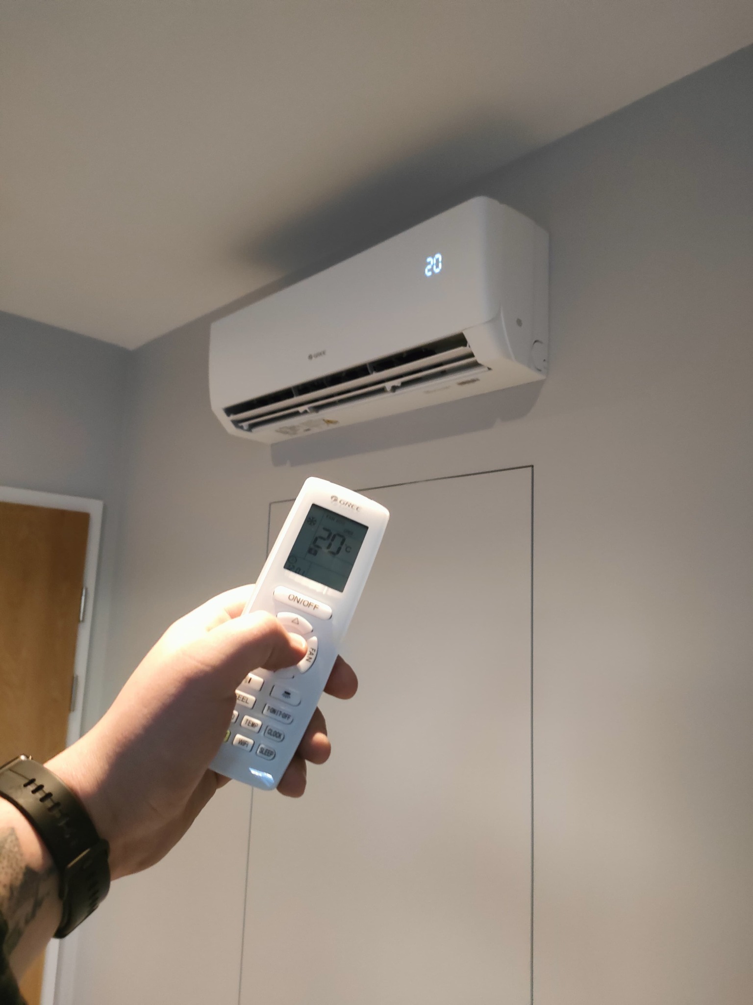 Biały klimatyzator Gree na ścianie, sterowany pilotem. Wyświetlacz pokazuje 20 stopni. Widoczna ręka z zegarkiem. Minimalistyczny design wnętrza.