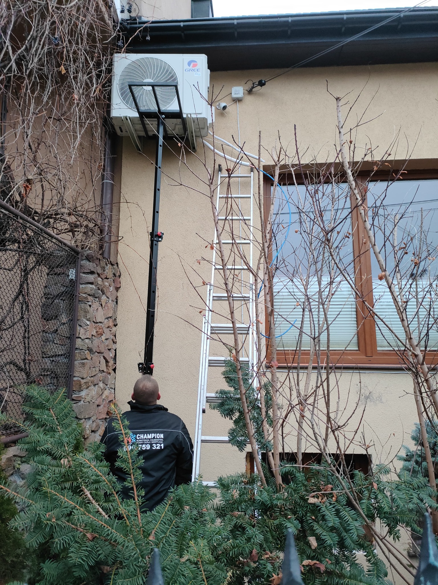 Montaż klimatyzatora Gree na elewacji budynku, widoczna drabina i pracownik w kurtce firmowej, zieleń w kadrze. Specyficzny kąt ujęcia.