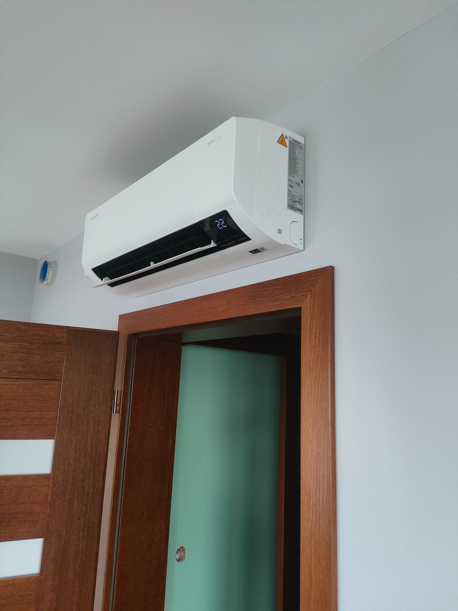 Biały klimatyzator Samsung WindFree zamontowany na ścianie nad drewnianymi drzwiami z szybami. Widoczny wyświetlacz z temperaturą 22 stopnie Celsjusza.