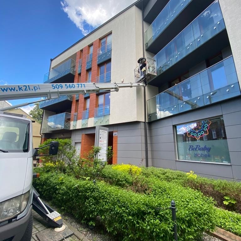 Montaż klimatyzacji na wysokości w budynku wielorodzinnym z użyciem podnośnika koszowego. Widoczny pracownik w trakcie pracy i fragment elewacji budynku.