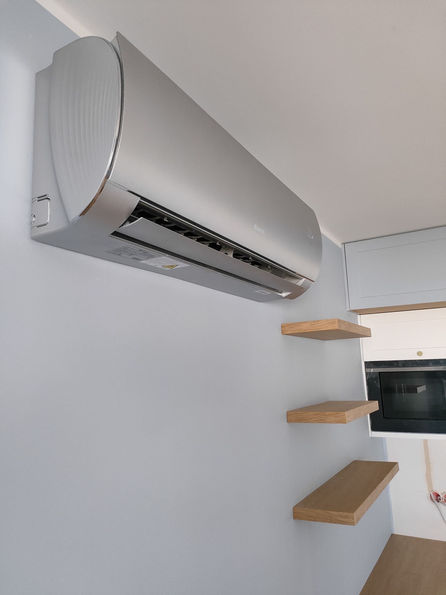Srebrny klimatyzator Gree zamontowany na szarej ścianie w pomieszczeniu z drewnianymi półkami i zabudową kuchenną. Widok z dołu, oświetlenie naturalne.