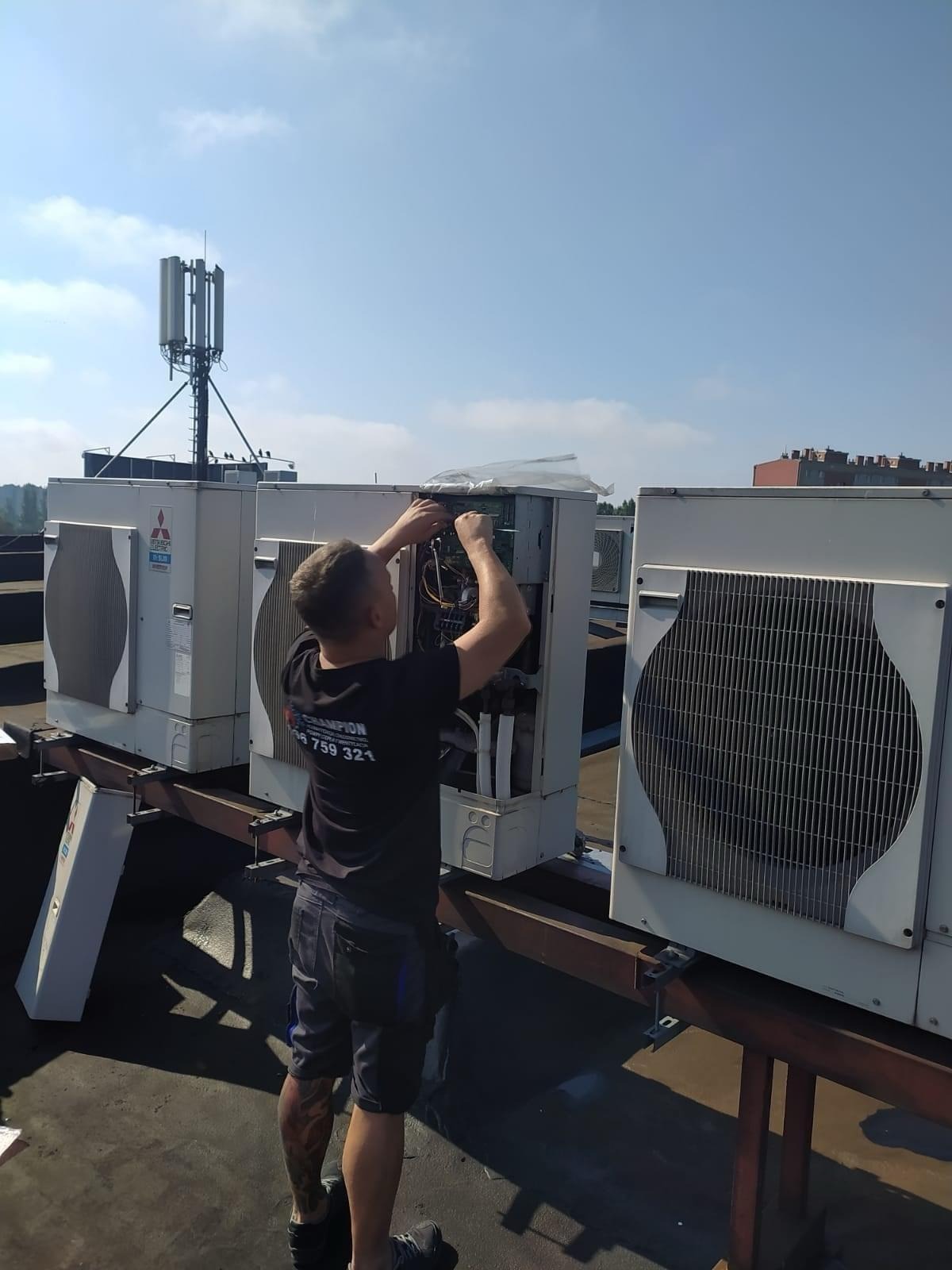 Instalator w czarnej koszulce serwisuje klimatyzator Mitsubishi na dachu budynku. Widoczna antena telekomunikacyjna w tle i błękitne niebo.