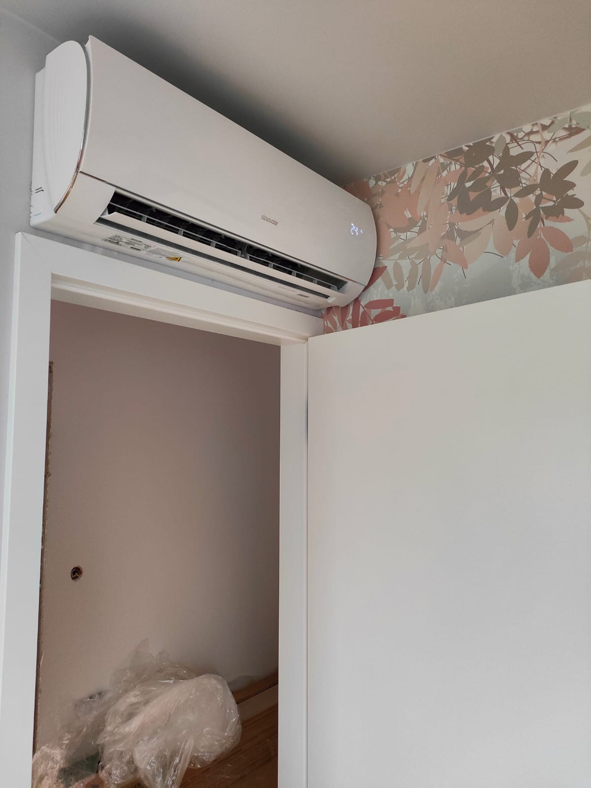 Biały klimatyzator Sinclair zamontowany nad futryną drzwi w pokoju z tapetą w liście, widoczny wyświetlacz temperatury 24 stopnie Celsjusza.