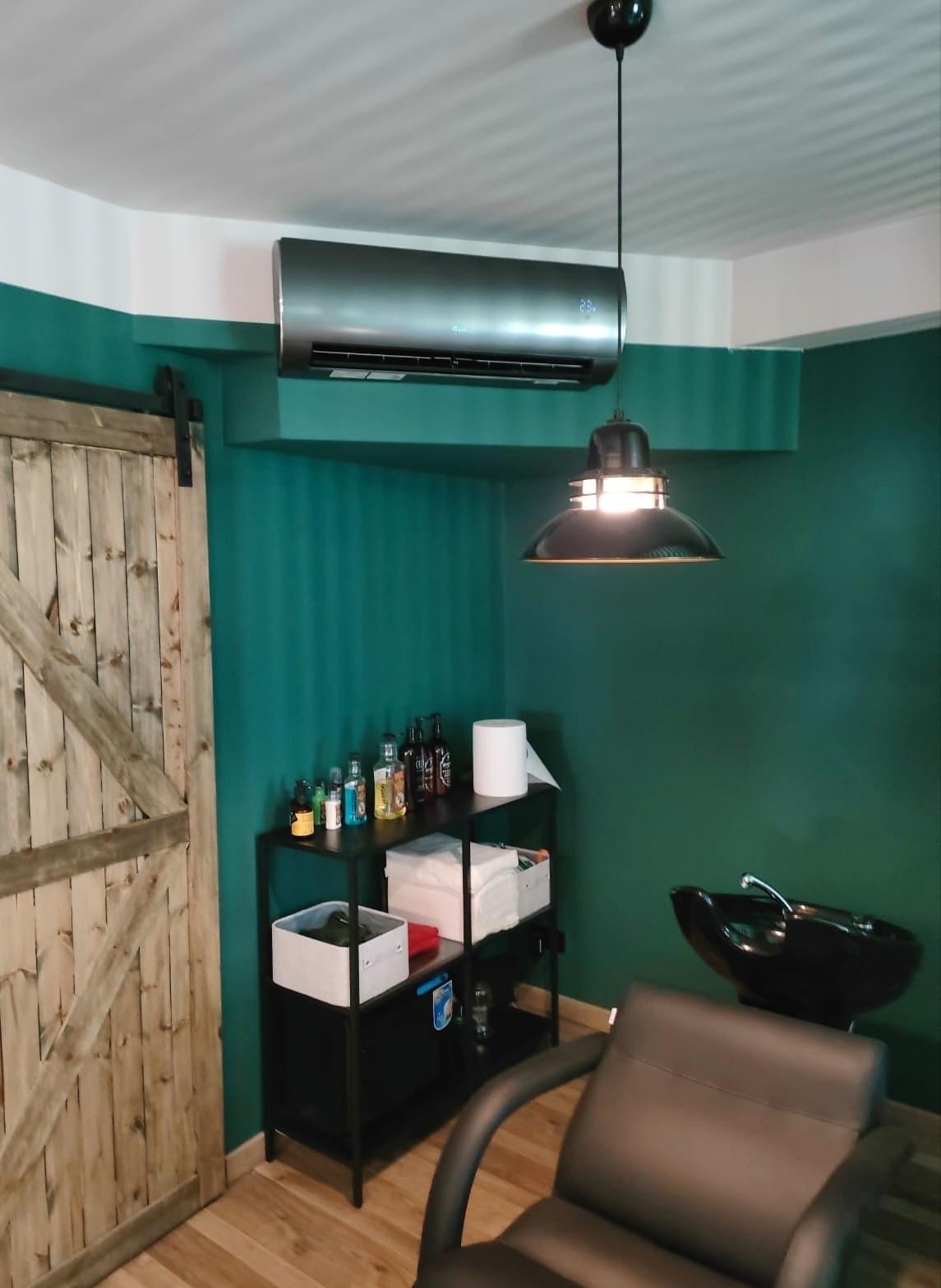 Wnętrze salonu fryzjerskiego z klimatyzatorem w kolorze grafitowym zamontowanym pod sufitem, drewnianymi drzwiami przesuwnymi, czarną lampą wiszącą, umywalką fryzjerską i półką z kosmetykami na tle...