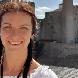 Uśmiechnięta kobieta w białej bluzce z falbankami i warkoczami na tle średniowiecznego zamku z powiewającymi flagami.