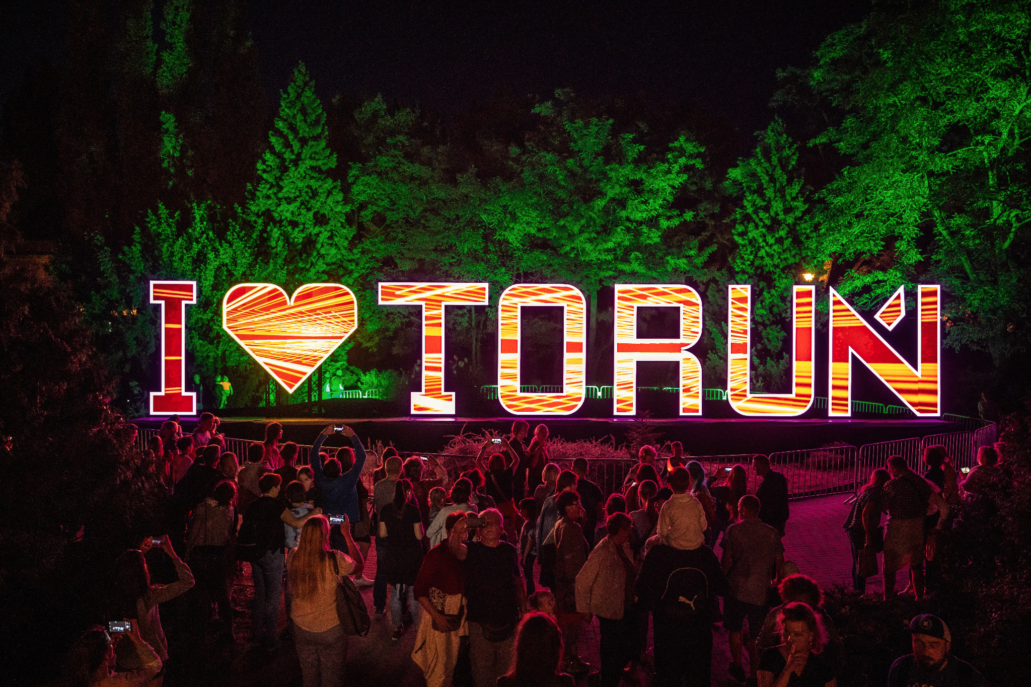 Tłum ludzi obserwuje podświetlaną instalację artystyczną 'I ❤️ TORUŃ' podczas Bella Skyway Festival, z zielonymi drzewami w tle.