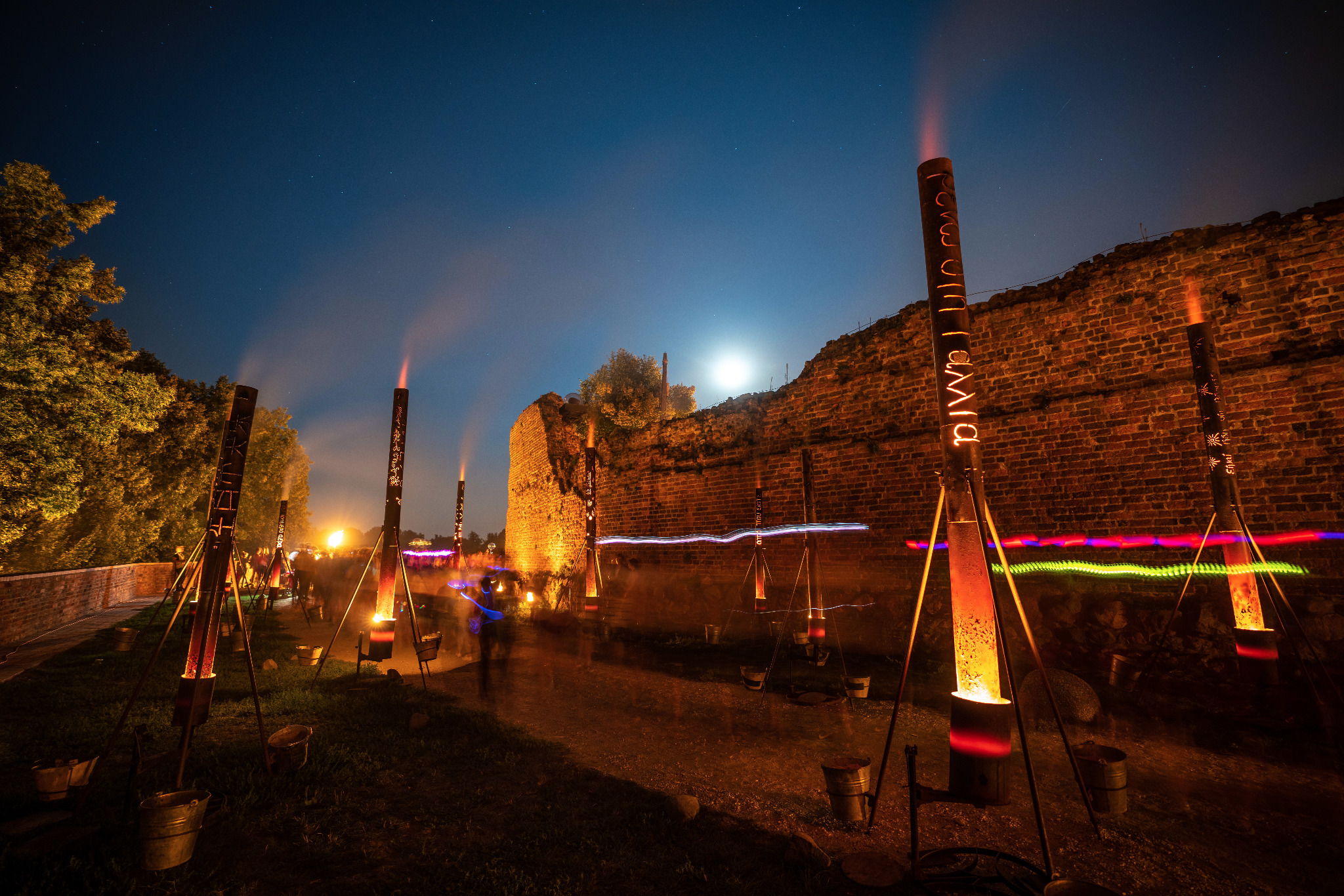 Nocna iluminacja Bella Skyway Festival w Toruniu: rząd metalowych słupów z płomieniami wewnątrz, ustawionych wzdłuż ceglanego muru, z widocznymi sylwetkami ludzi w tle i księżycem na niebie.