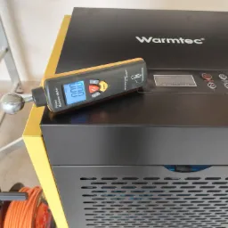 Czarny osuszacz powietrza marki Warmtec z żółtymi elementami obudowy, na którym leży miernik wilgotności pinless z wyświetlaczem LCD pokazującym wartość 0.00, obok widoczne zwinięte kable.