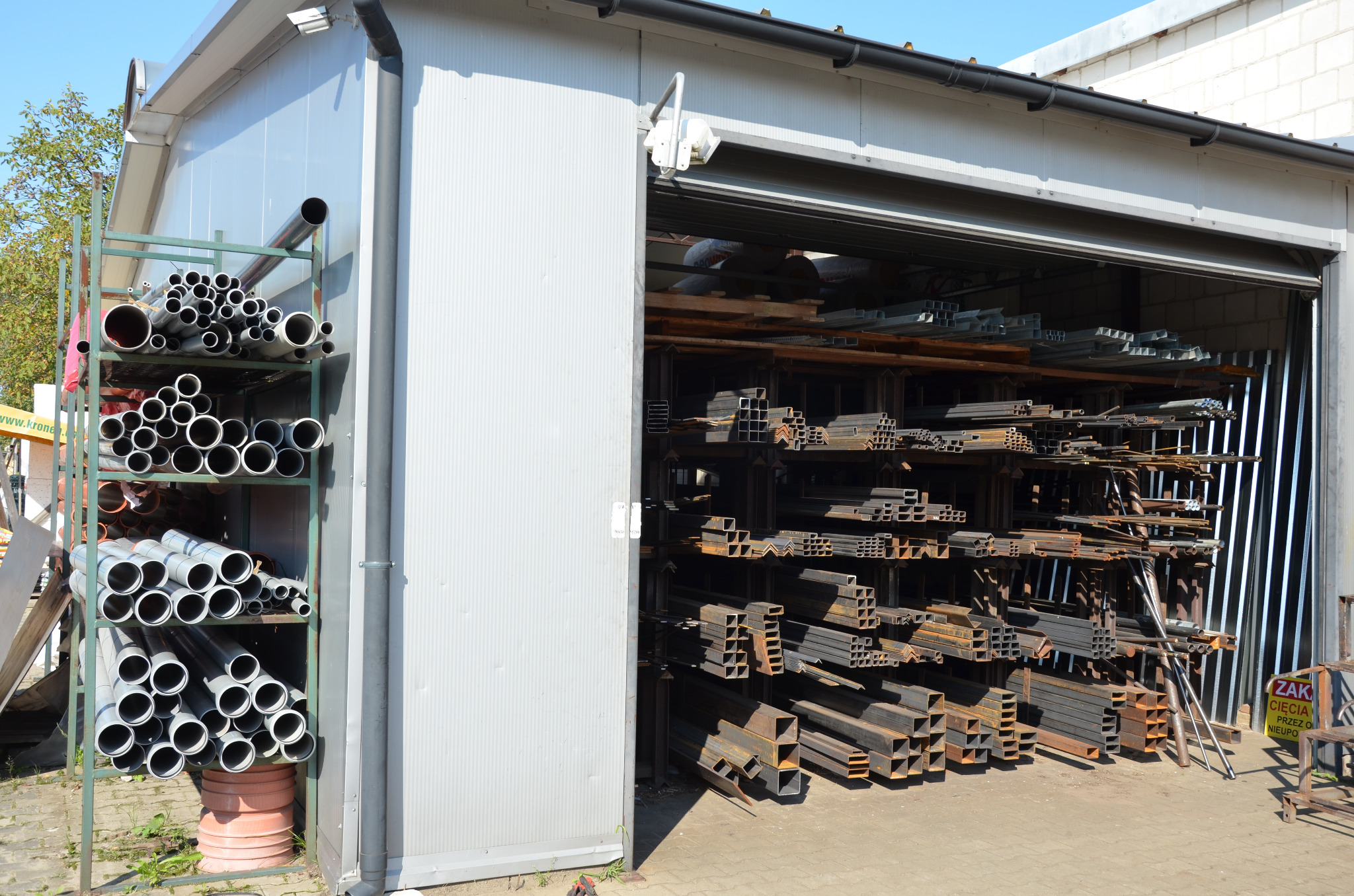 Otwarty magazyn z półkami pełnymi rur PCV i metalowych profili o różnych przekrojach, ułożonych warstwami.