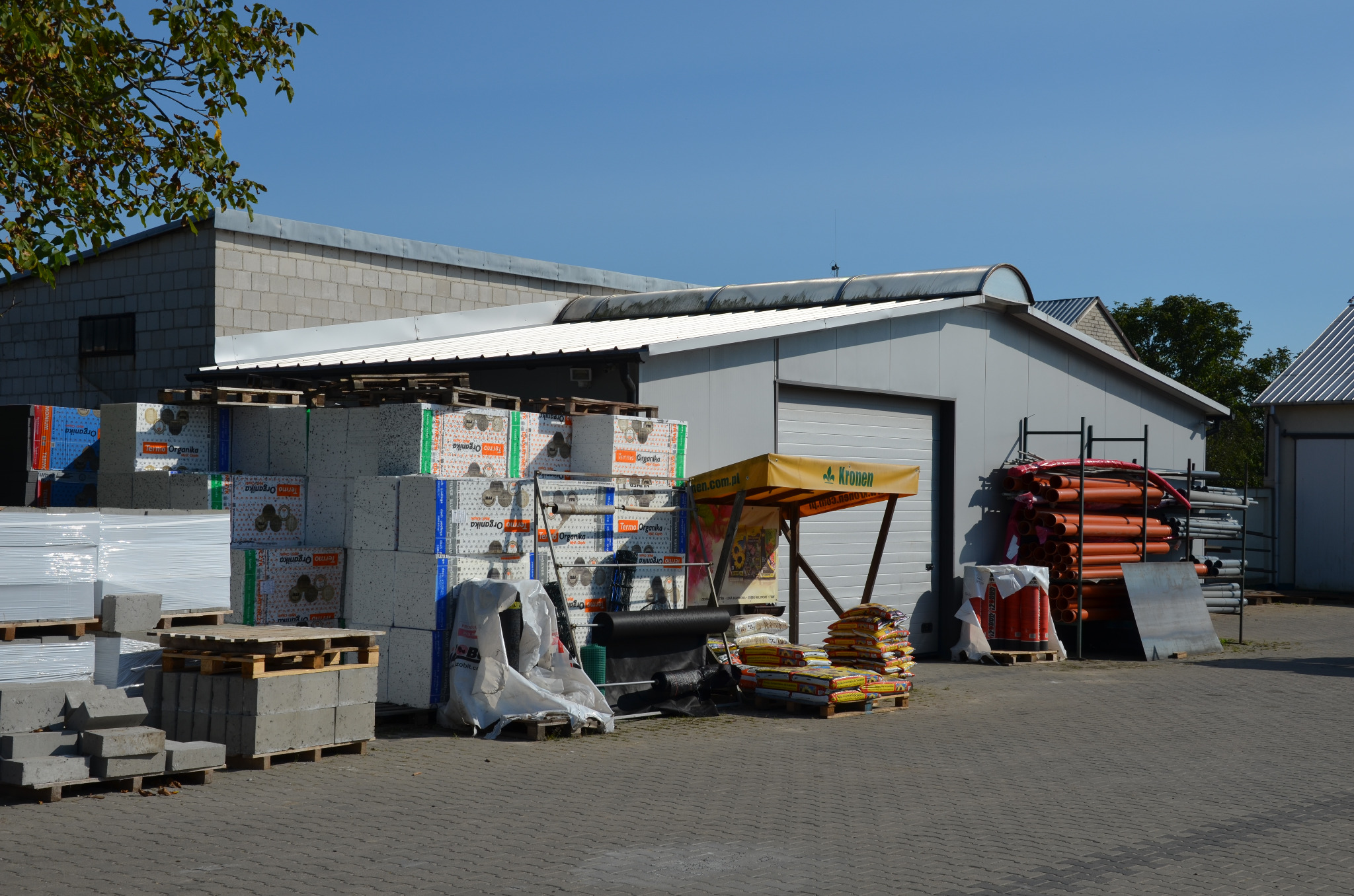 Składowisko materiałów budowlanych: palety z bloczkami betonowymi i styropianem Termo Organika, rury PCV oraz worki z zaprawą pod żółtą markizą reklamową Kronen na tle magazynu z bramą rolowaną.