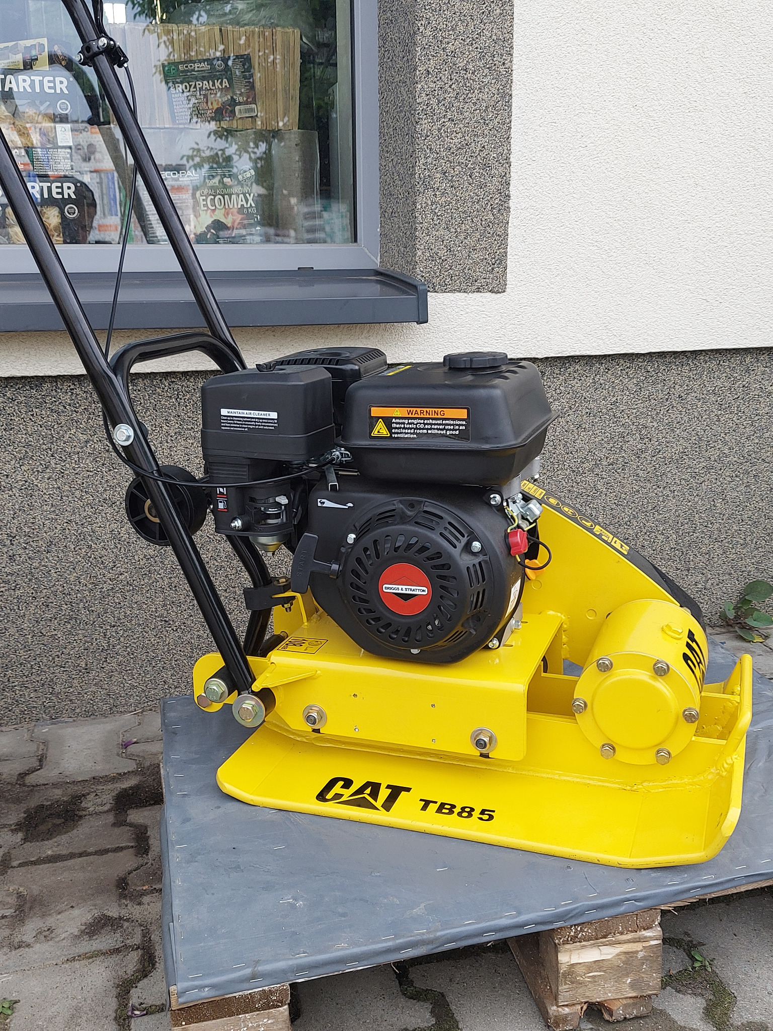 Żółta zagęszczarka gruntu CAT TB85 na drewnianej palecie przed budynkiem z szarą elewacją, widoczna etykieta ostrzegawcza i silnik Briggs & Stratton.
