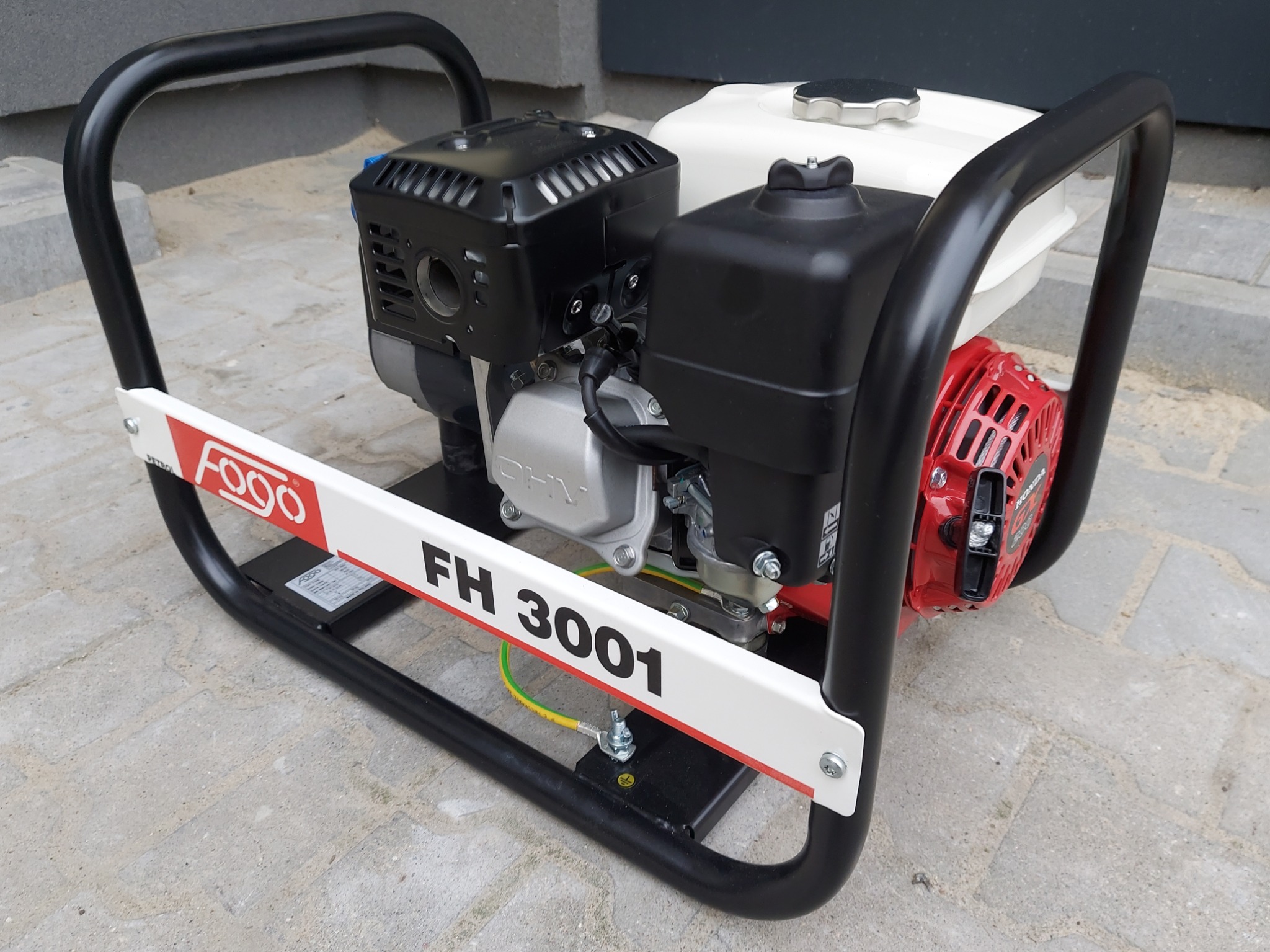 Agregat prądotwórczy Fogo FH 3001 z widocznym uziemieniem na białej ramie, na tle szarej kostki brukowej.