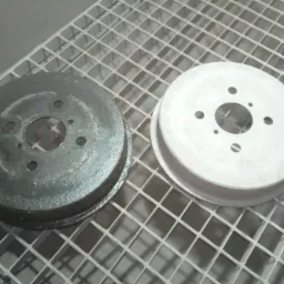 Dwa bębny hamulcowe na metalowej kratce: jeden czarny, pokryty resztkami rdzy, drugi po piaskowaniu, czysty, o jasnoszarym odcieniu.