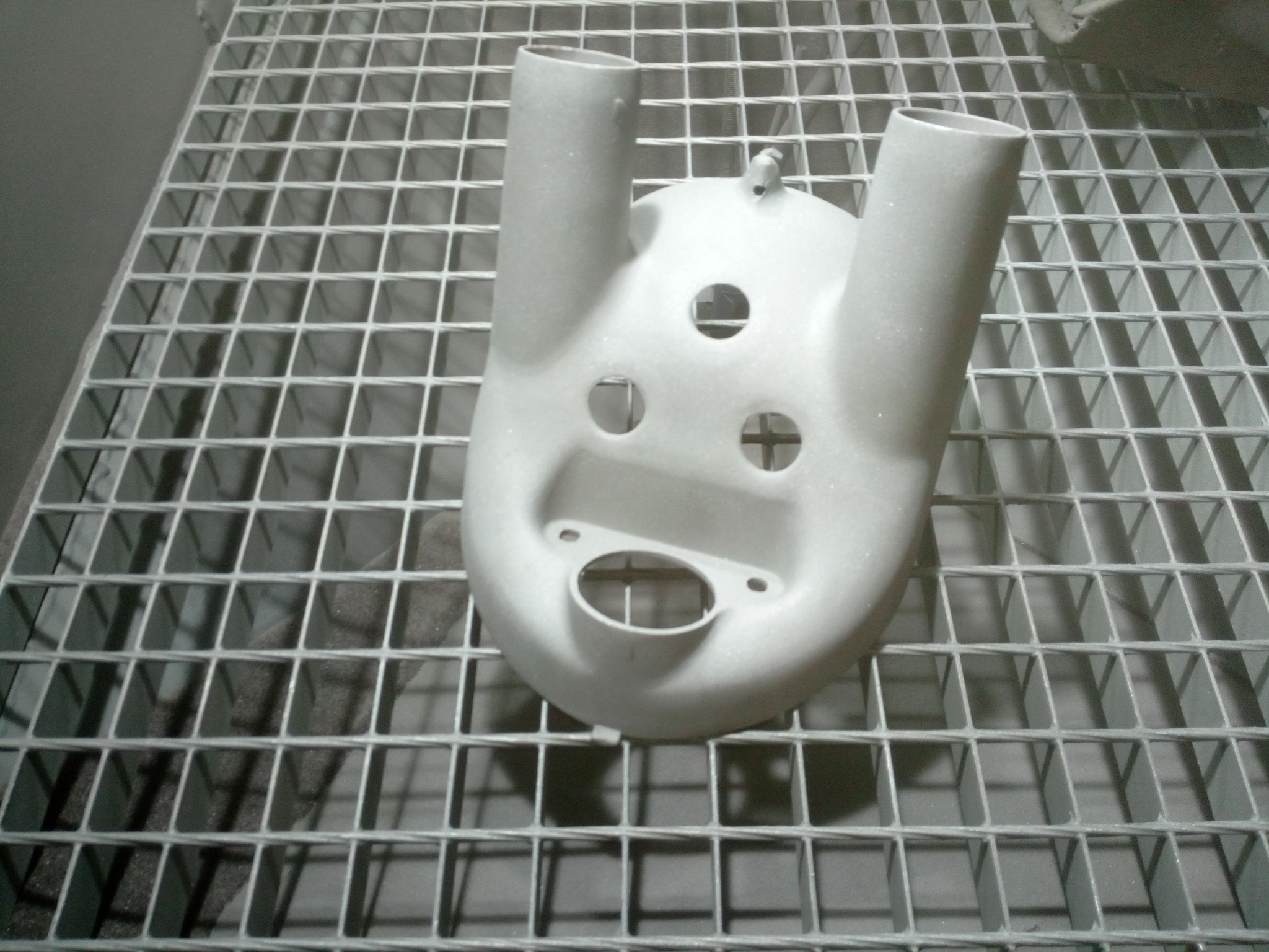 Metalowa obudowa z otworami i dwoma wystającymi rurami, leżąca na metalowej kratce, po obróbce strumieniowo-ściernej.