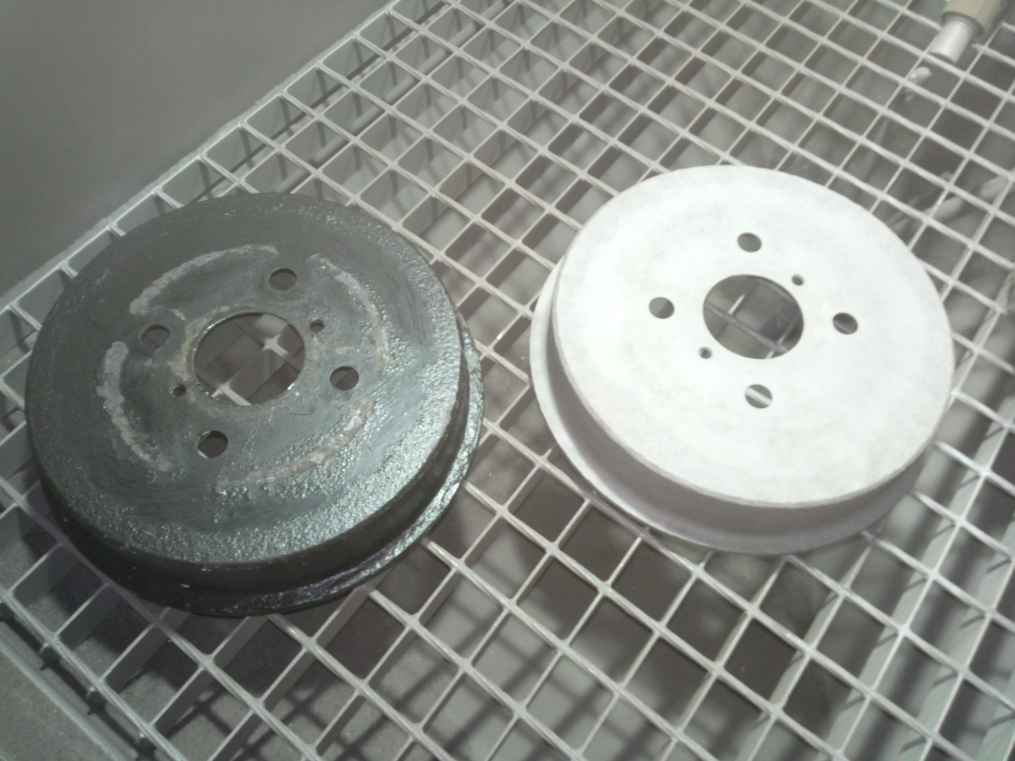 Dwa bębny hamulcowe na metalowej kratce: jeden czarny, pokryty resztkami rdzy, drugi po piaskowaniu, czysty, o jasnoszarym odcieniu.