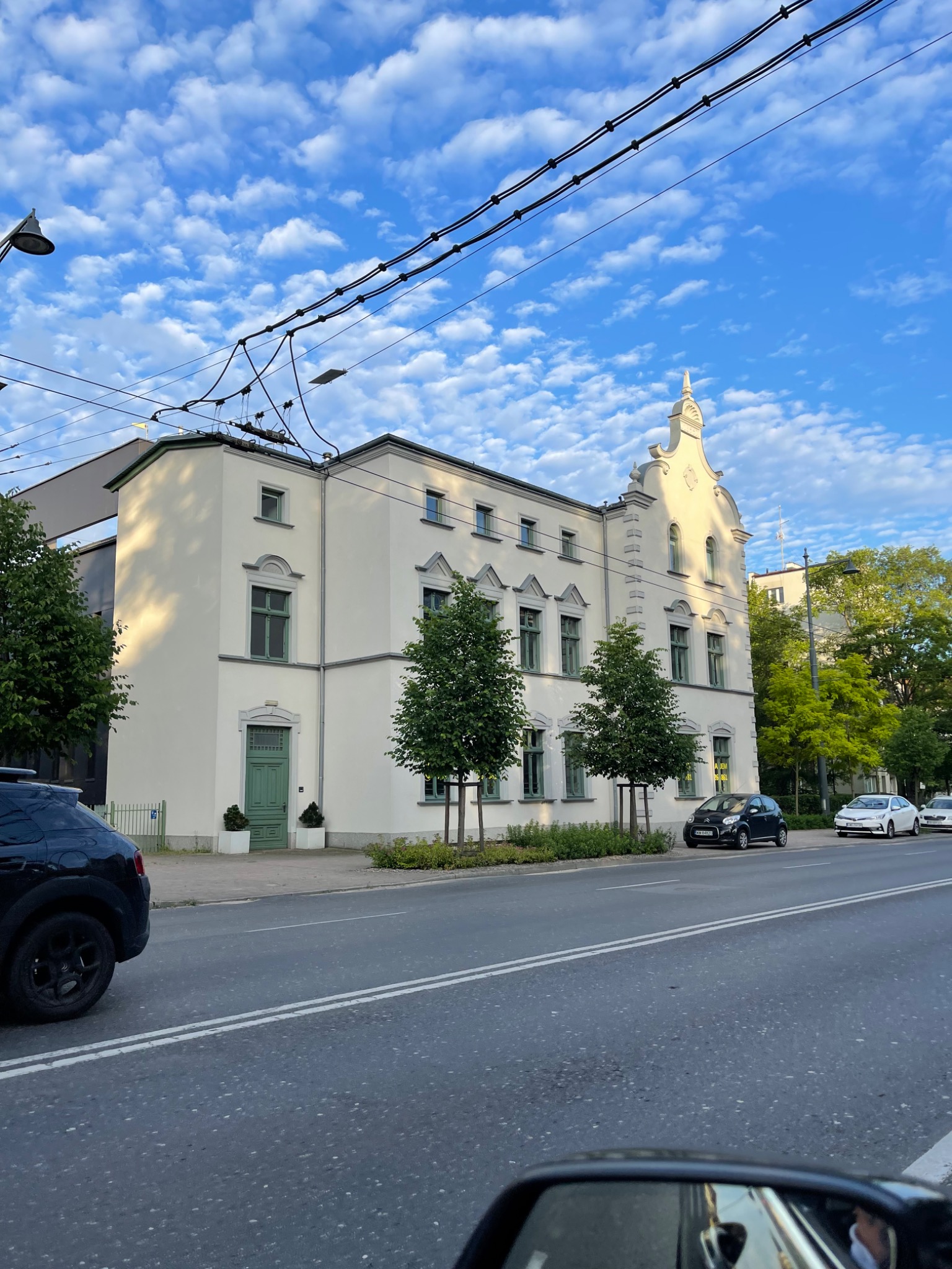 Elewacja odnowionego, trzypiętrowego budynku usługowego w stylu klasycystycznym z zielonymi oknami i drzwiami, widziana z perspektywy ulicy w słoneczny dzień z błękitnym niebem i obłokami.