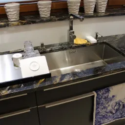 Błyszczący, stalowy zlew jednokomorowy w blacie z ciemnego granitu z wyraźnym użyleniem, bateria kuchenna o kwadratowym przekroju, na blacie białe doniczki z roślinami i akcesoria kuchenne,...