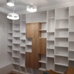 Biała, zabudowana półka na książki z trzema drewnianymi szafkami pośrodku, oświetlona nowoczesnymi lampami sufitowymi.