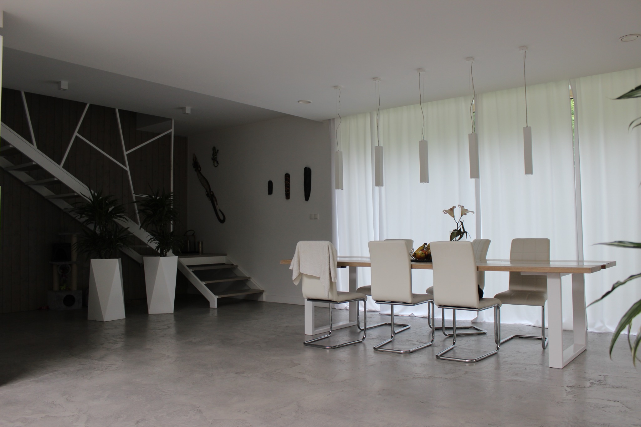 Jasne, przestronne wnętrze z betonową podłogą, drewnianym stołem otoczonym krzesłami z jasnej skóry i metalu, minimalistycznymi lampami wiszącymi oraz schodami z geometryczną balustradą.